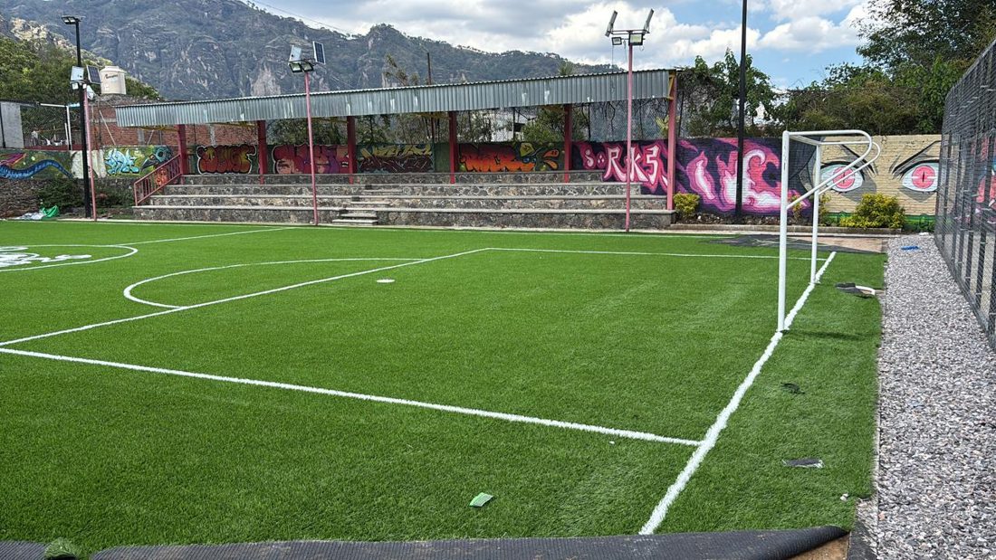 Cancha de fútbol de pasto sintético con gradas techadas, un mural de grafiti y montañas al fondo